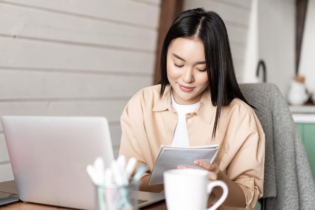 Aziatische studente doet huiswerk jonge vrouw die aantekeningen maakt terwijl ze online webinar bijwoont en aan la...