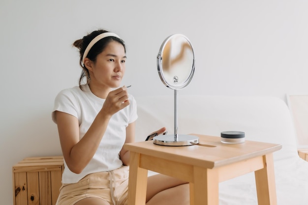 Foto aziatische thaise vrouw met potlood en het tekenen van wenkbrauwen zit op een witte bank voor de spiegel in