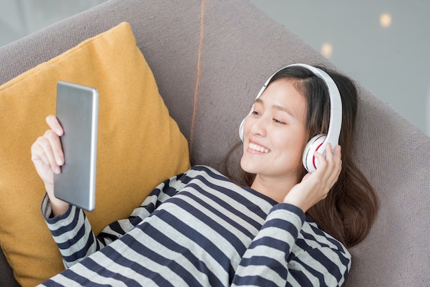 Aziatische tiener meisje dragen hoofdtelefoon luisteren muziek spelen met tablet pc lag op de bank