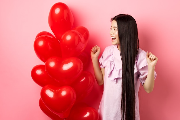Aziatische tienermeisje met lang haar, juichen van Valentijnsdag romantisch cadeau, logo kijken en glimlachen gelukkig, springen van vreugde in de buurt van liefhebbers cadeau hart ballonnen, roze achtergrond.