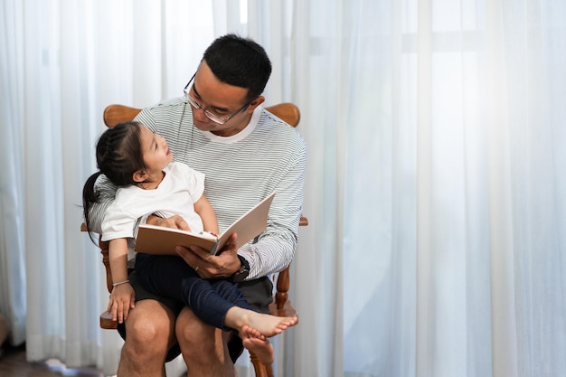 Aziatische vader die verhalenboek leest aan dochter in het familieconcept van de woonkamer en ontwikkeling in de vroege kinderjaren