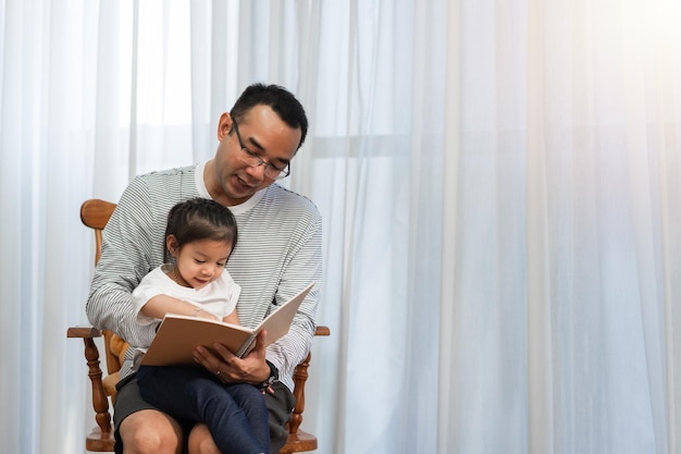 Aziatische vader die verhalenboek leest aan dochter in het familieconcept van de woonkamer en ontwikkeling in de vroege kinderjaren