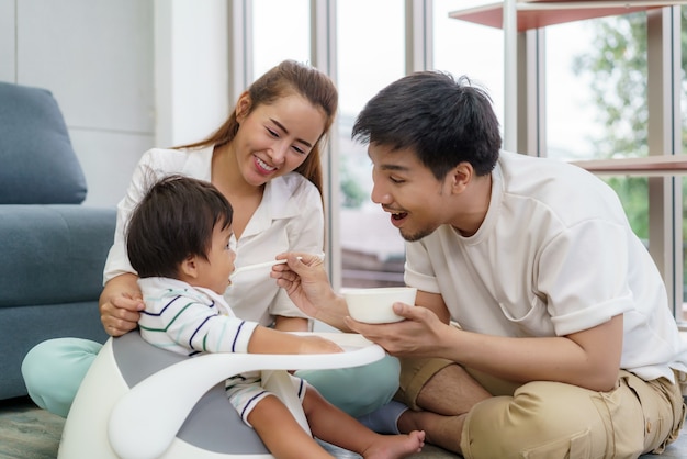 Aziatische vader die zijn 6 maanden oude babyjongen voedt met vast voedsel met lepel en moeder die dichtbij haar zoon zit toe te juichen om voedsel in de woonkamer thuis te eten.