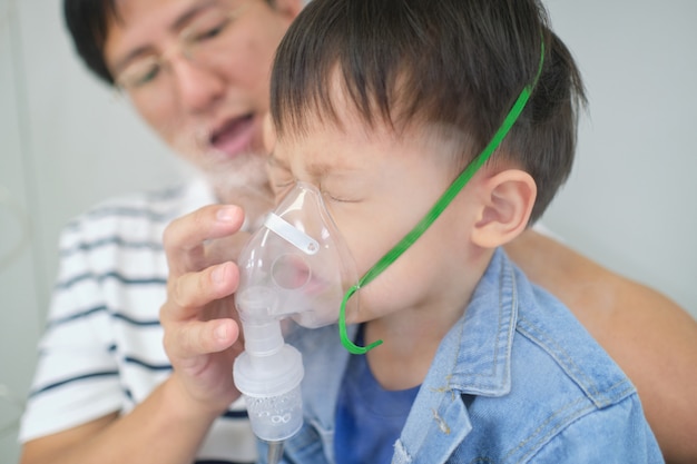 Aziatische vader die zijn peuterzoon helpt met inhalatietherapie door het masker van inhalator. Ziek klein kind met ademhalingsproblemen met zuurstofmasker ademt door verstuiver