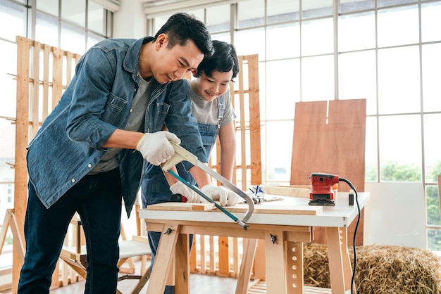 Aziatische vader en zoon werken als houtbewerker of timmerman Vader leert zijn zoon een houten plank te zagen met een ijzerzaag zorgvuldig samen met teamwerk Ambachtsman timmerwerk thuis werken workshop studio