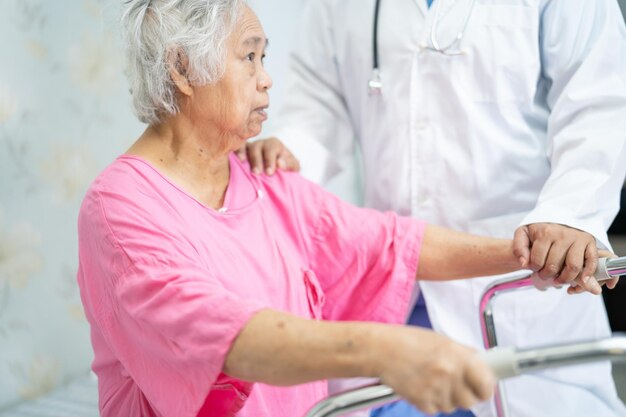 Aziatische verpleegster fysiotherapeut arts zorg, hulp en ondersteuning senior of oudere oude dame vrouw patiënt lopen met rollator op ziekenhuisafdeling, gezond sterk medisch concept.