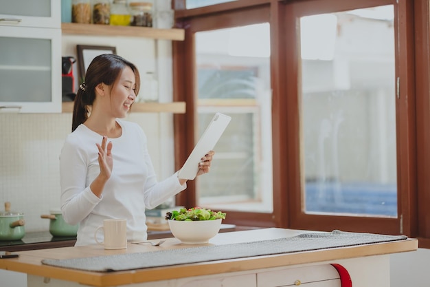 Aziatische vrouw begroet haar vrienden via digitale tablet in de keuken voordat ze saladegroenten eet