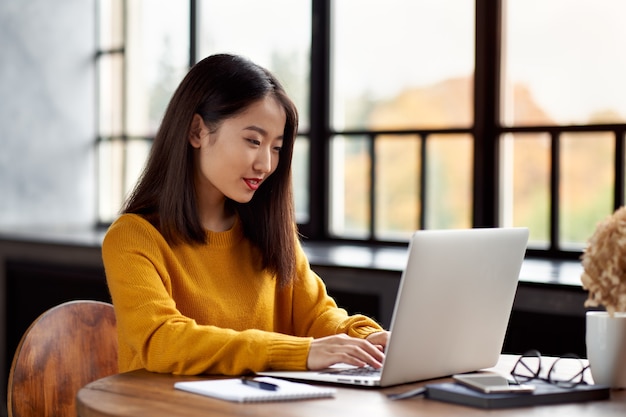 Aziatische vrouw die aan laptop thuis of in koffie werkt. Jonge dame in felgele trui