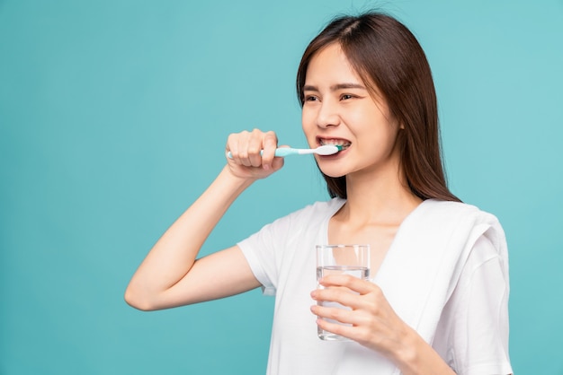 Aziatische vrouw die beugels draagt met tanden poetsen en waterglas vasthoudt, handdoek op de schouder op blauwe achtergrond, Concept mondhygiëne en gezondheidszorg.
