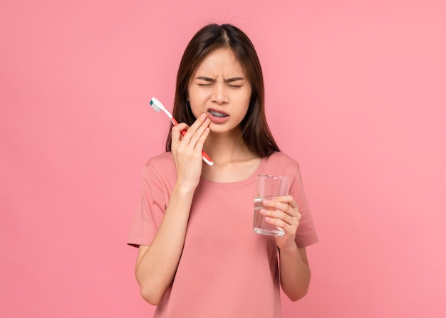 Aziatische vrouw die beugels draagt met tandenpoetsen met gevoelige tanden en waterglas vasthoudt op roze achtergrond, Concept mondhygiëne en gezondheidszorg.