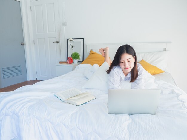 Aziatische vrouw die computer met behulp van en boek op bed in de ochtend leest