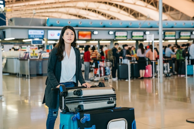 Aziatische vrouw die door de luchthavenbalie loopt en naar de terminal duwt met handbagage voor vliegreizen. mensen in de rij op de achtergrond wachten om in te checken. jonge vrouwelijke werknemer.