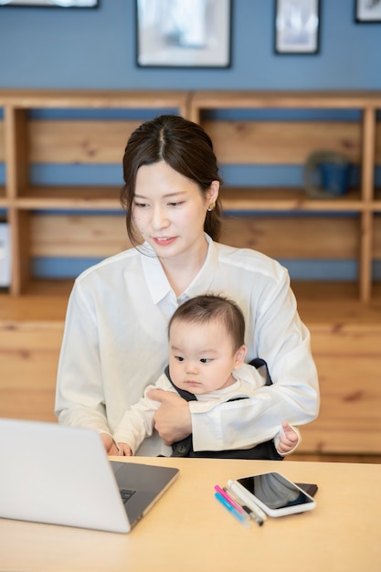 Aziatische vrouw die een baby houdt en laptop binnen in werking stelt