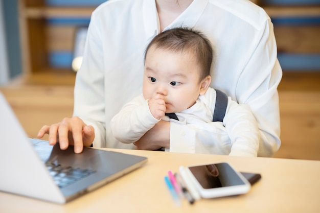 Aziatische vrouw die een baby houdt en laptop binnen in werking stelt