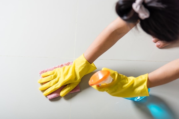 Aziatische vrouw die een gele rubberen handschoen draagt met een doek en een schoonmaakspray die de vloer thuis schoonmaakt