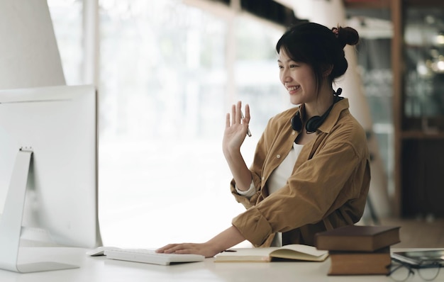 Aziatische vrouw die een koptelefoon op haar nek draagt met behulp van laptop en vrienden begroet tijdens een videogesprek thuis