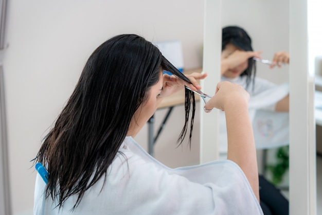 Aziatische vrouw die haar haar zelf knipt met een knipschaar, ze blijven thuis en schuilen op hun plaats tijdens de isolatie van het huis tegen nieuw coronavirus of covid-19