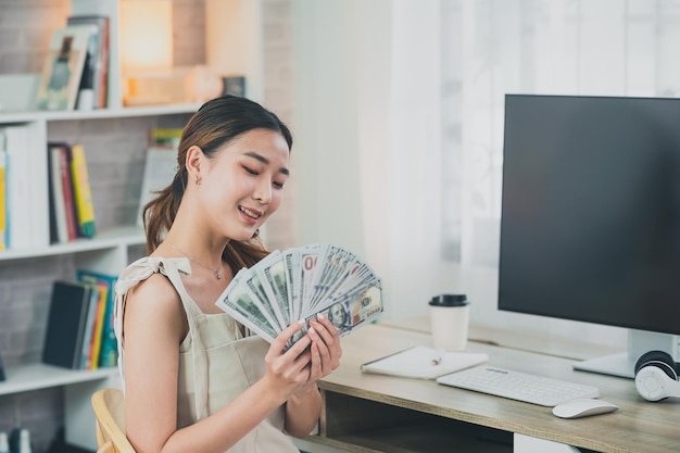 Aziatische vrouw die lacht en een dollarbiljet vasthoudt op een houten tafelbureau in de woonkamer thuis