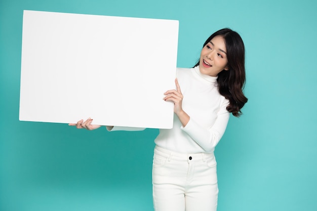 Aziatische vrouw die leeg wit reclamebord toont en houdt dat op groene achtergrond wordt geïsoleerd