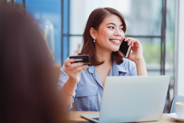 Aziatische vrouw die mobiele telefoon met creditcard en laptop computer voor het winkelen online betaling in koffiewinkel koffie met vrienden met behulp van