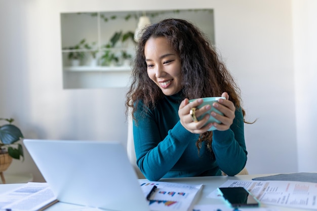 Aziatische vrouw die op laptop computer werkt die lacht Vrouw die vanuit huis werkt op laptop in modern appartement Trendy vrouw die op laptop werkt en koffie drinkt