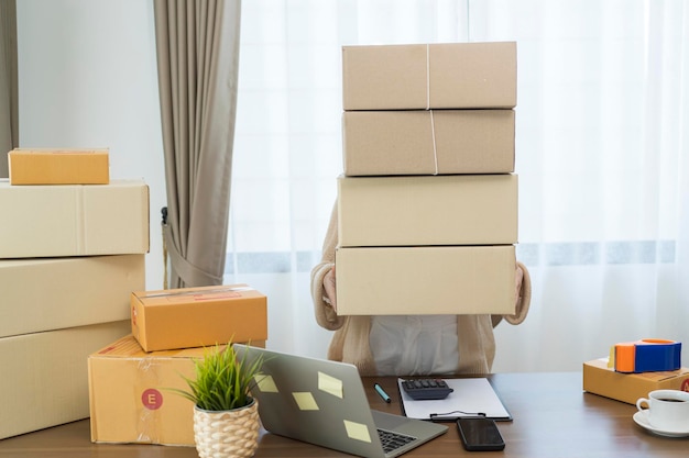 Aziatische vrouw die pakketdozen vasthoudt en bestellingen van laptop controleert. Ze heeft een online winkel die ze inpakt en verzendt via een particulier transportbedrijf