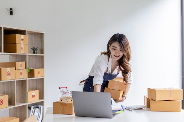 Aziatische vrouw die pakketdozen vasthoudt en bestellingen van laptop controleert, ze is eigenaar van een online winkel, ze verpakt en verzendt via een particulier transportbedrijf. Online verkoop en online winkelconcepten.