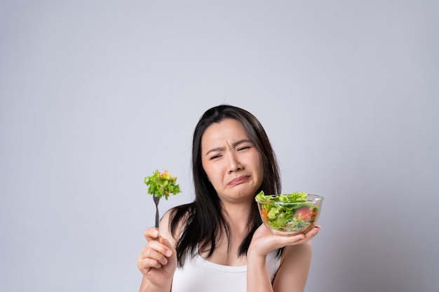 Aziatische vrouw die salade voor dieet probeert te eten dat over witte muur wordt geïsoleerd. Gezonde levensstijl met schoon voedselconcept.