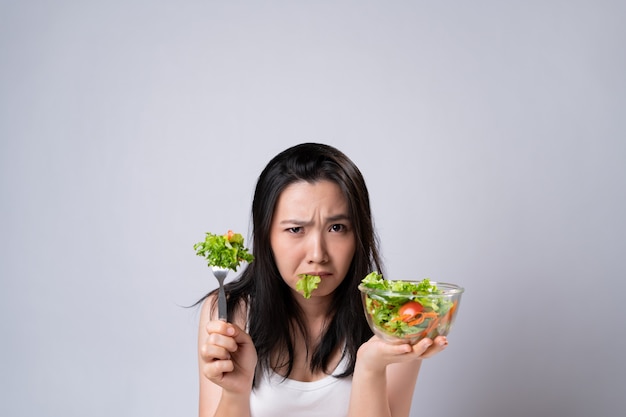 Aziatische vrouw die salade voor dieet probeert te eten dat over witte muur wordt geïsoleerd. Gezonde levensstijl met schoon voedselconcept.