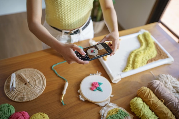 Aziatische vrouw die smartphone foto neemt Punch naald telefoon posten op sociale netwerken in studio werkplaats ontwerper werkplek Handgemaakt ambachtelijk project DIY borduren