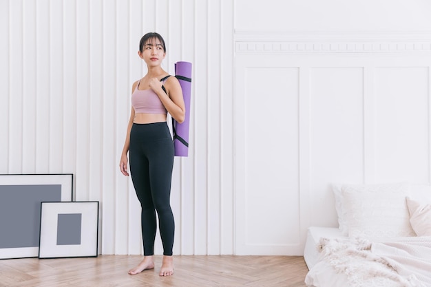 Aziatische vrouw die sportkleding en yogabroek draagt die een opgerolde yogamat dragen die zich in een slaapkamer bevindt