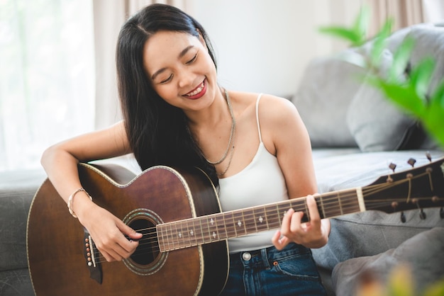 Aziatische vrouw die thuis muziek speelt met gitaar, jonge vrouwelijke gitarist muzikant levensstijl met akoestisch kunstinstrument zit om te spelen en een lied te zingen dat geluid maakt in hobby in de huiskamer