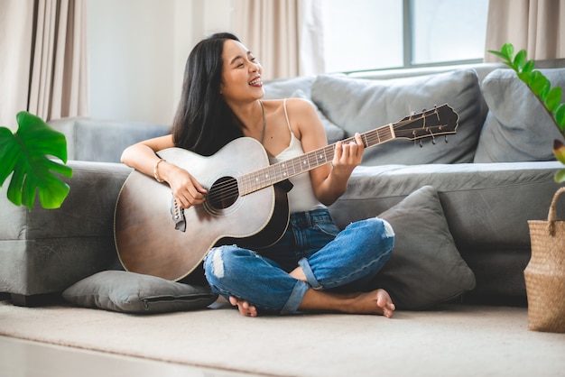 Aziatische vrouw die thuis muziek speelt met gitaar, jonge vrouwelijke gitarist muzikant levensstijl met akoestisch kunstinstrument zit om te spelen en een lied te zingen dat geluid maakt in hobby in de huiskamer