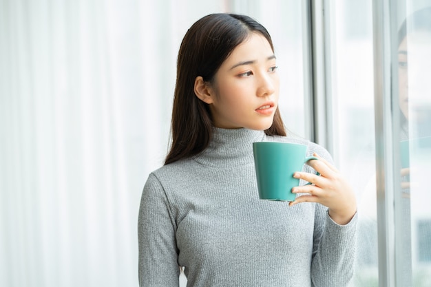 Aziatische vrouw die tijdens de pauze koffie drinkt bij het raam