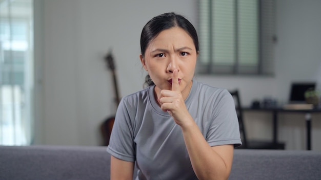 Aziatische vrouw die wijsvinger op de lippen legt, wat betekent dat ze geheim moet houden en naar de camera kijkt die op de bank in de woonkamer thuis zit