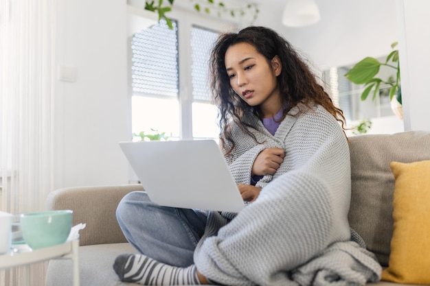 Aziatische vrouw gewikkeld in een gezellige deken zittend op de bank en bezig met haar laptop