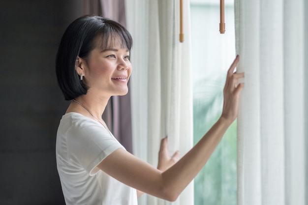 Aziatische vrouw glimlach en kijken naar buiten met gordijn op een kamer