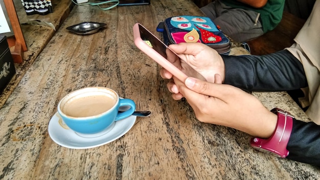 Aziatische vrouw hand met een mobiele telefoon met een kopje cappuccino op houten tafel