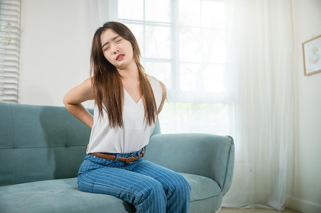 Aziatische vrouw houdt haar onderrug vast en voelt pijnlijke taille pijn massage in de woonkamer, mooie jonge vrouw raakt rugpijn aan terwijl ze op de bank zit, ze lijdt aan rugpijn thuis, medische gezondheidszorg