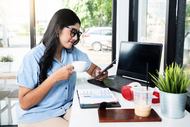 Aziatische vrouw in bril met een laptop en notebook op tafel met een kopje koffie tijdens het gebruik van een mobiele telefoon