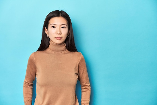 Aziatische vrouw in bruine turtleneck kijkt naar camera blauwe achtergrond