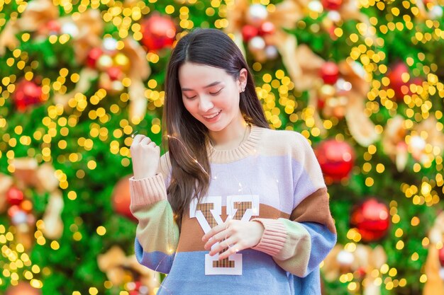 Aziatische vrouw in een kleurrijke trui staat vrolijk voor de kerstboom