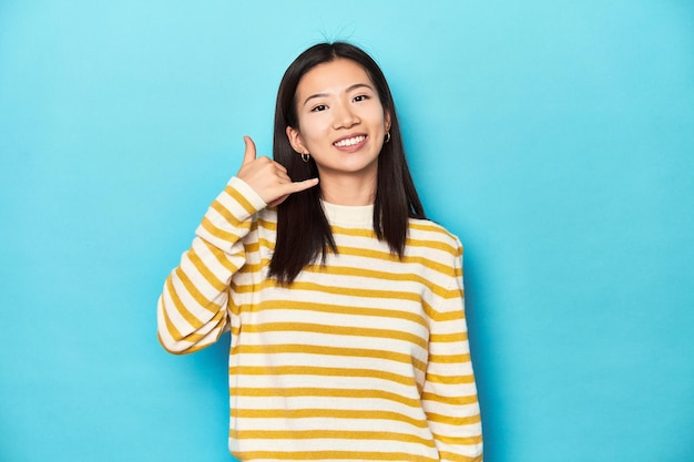 Aziatische vrouw in gestreepte gele trui die een mobiele telefoongesprek met vingers toont