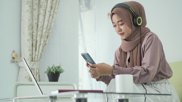 Aziatische vrouw in hijab op zoek naar inspiratie op smartphone terwijl ze luistert via een koptelefoon