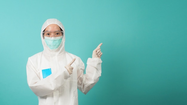 Aziatische vrouw in PPE-pak draagt een gezichtsmasker en wijst met de vinger omhoog op een groene of Tiffany Blue-achtergrond.