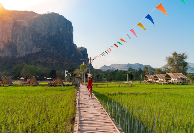 Aziatische vrouw in rode jurk met hoed staande op houten brug over groene rijstveld kijken naar grote bergzicht (Phu Pha Man) in Khonkaen, Thailand, landschap van verse rijst boerderij en heuvel