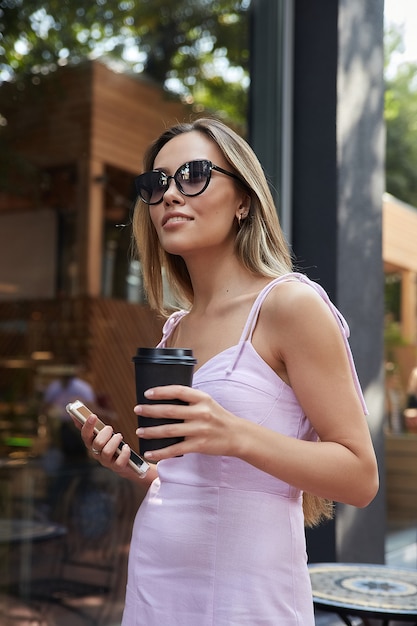 Aziatische vrouw in roze jurk die buiten staat met smartphone en kopje koffie