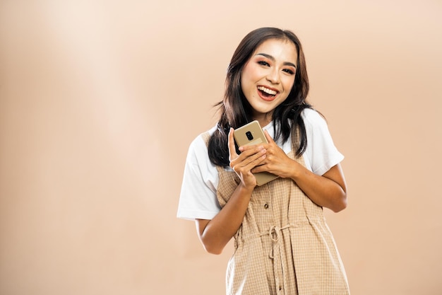 Aziatische vrouw in vrijetijdskleding die met een glimlach staat en de telefoon vasthoudt met haar beide handen op isola