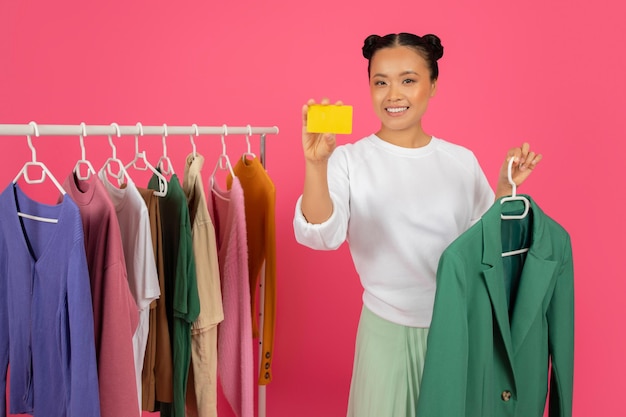 Aziatische vrouw met creditcard en blazer terwijl ze in de buurt van kledingrek staat