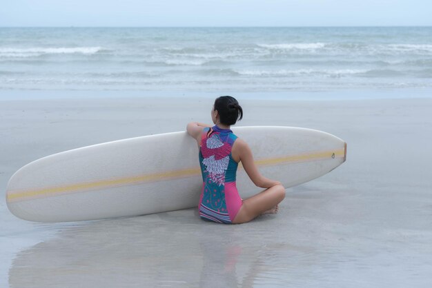 Aziatische vrouw met een surfplank naast me zit op het zand en kijkt naar de golven in de zee.